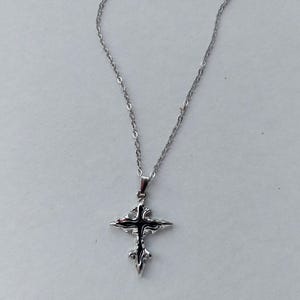 May include: A silver pendant necklace with a decorative cross. The cross has an ornate, detailed design with a dark centre. The necklace chain is delicate and hangs against a white background.