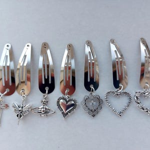 May include: A collection of silver-coloured metal hair clips, each with a different charm. Charms include a cross, a winged insect, a planet, a heart, a heart with a cross, a heart outline, and a barbed wire heart. Clips are in a row.