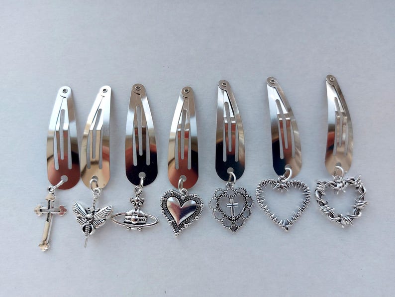 May include: A collection of silver-coloured metal hair clips, each with a different charm. Charms include a cross, a winged insect, a planet, a heart, a heart with a cross, a heart outline, and a barbed wire heart. Clips are in a row.