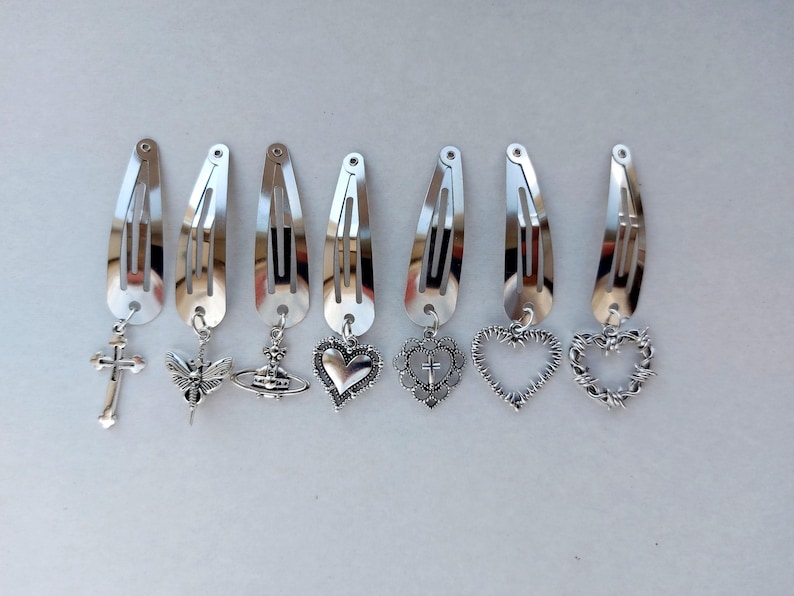 May include: Seven silver hair clips with decorative charms. The charms include a cross, a winged insect, a globe, a heart, a heart with a cross, a heart outline, and a heart with barbed wire. The clips are displayed in a line.