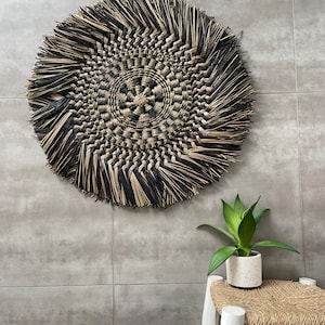 Puede incluir: Un gran tapiz de pared redondo hecho de fibras naturales tejidas en negro y marrón. El tapiz tiene un borde de flecos y un patrón circular en el centro. El tapiz está montado en una pared gris.