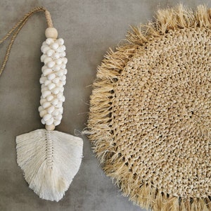 May include: A white tassel with a string of white beads hanging from it. The tassel is on a grey background next to a round, woven straw placemat with a fringe border.