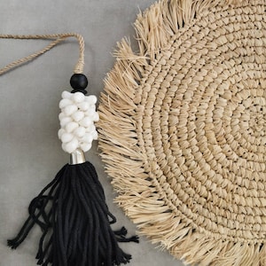 Può includere: Un fiocco nero con una perla di conchiglia bianca e una perla di legno nero. Il fiocco è appeso a una corda di fibra naturale. Il fiocco è su un sottopentola di paglia intrecciata con un bordo a frangia.