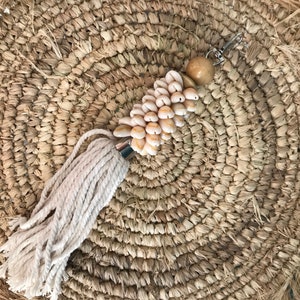 Op de afbeelding: Een witte kwast sleutelhanger met een cluster van kleine schelpen en een houten kraal. De sleutelhanger ligt op een geweven stro achtergrond.