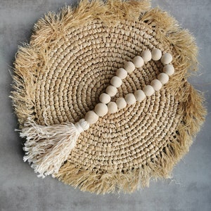 Puede incluir: Una guirnalda de cuentas de madera beige con un borla blanca colgando de un mantel individual redondo de paja trenzada con un borde de flecos.