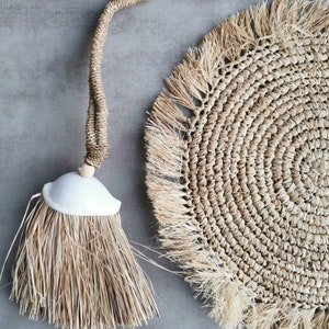 以下が含まれることがあります： 天然繊維製のホームデコレーションアイテム2点セット。フリンジの縁取りが施された丸い織りのプレースマットと、白いセラミックのトップが付いた天然繊維製のタッセル。