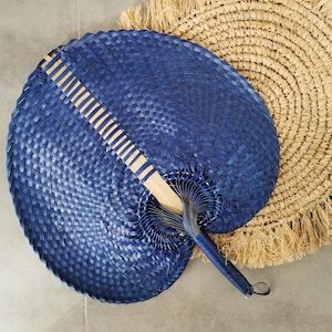 May include: A large, round, navy blue hand fan with a woven design. The fan has a wooden handle and a loop for hanging. A natural woven placemat with fringe is in the background.