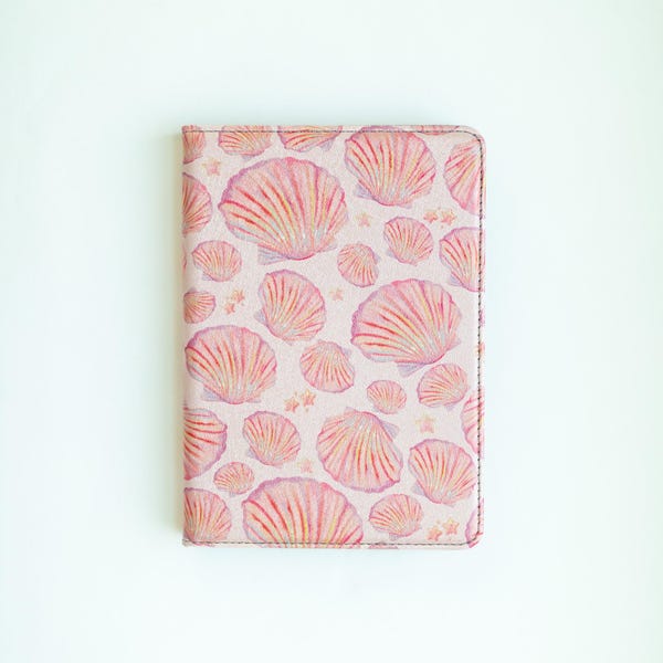May include: A pink notebook cover featuring a repeating pattern of watercolor seashells in shades of pink and peach. The cover has a stitched border and small star accents. The design evokes a beach or coastal theme.
