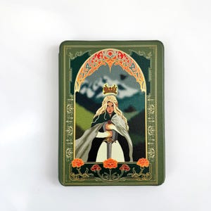 May include: A green rectangular case with a fantasy illustration. The artwork shows a blonde woman wearing a crown and holding a sword, set against a mountain backdrop. The design includes decorative floral and arch elements.