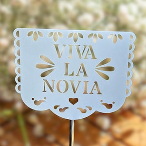 May include: A light blue paper banner with the words "VIVA LA NOVIA" cut out. The banner has a scalloped edge and is mounted on a thin metal rod. The background is blurred, with a soft, floral appearance.