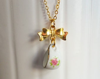 Puede incluir: Un collar dorado con un pequeño colgante de taza de té blanca con un diseño de rosa rosa. La taza de té cuelga de un dije de lazo dorado. El collar se muestra sobre un fondo blanco.