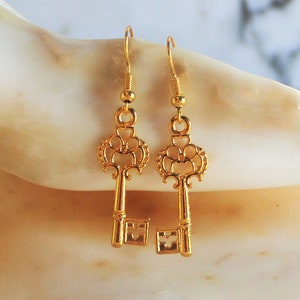May include: A pair of gold key-shaped earrings with intricate details. The earrings are hanging from a white seashell.