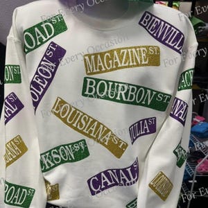May include: White sweatshirt featuring street signs in green, gold, and purple. The signs include "Bourbon St", "Canal St", and "Magazine St". A matching glitter top hat is positioned above the sweatshirt.