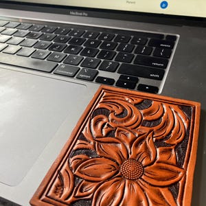 May include: A brown leather coaster with a tooled floral design. The coaster features a large flower in the center with intricate details and smaller floral patterns surrounding it.