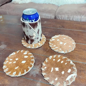 May include: Four brown and white cowhide coasters with white stitching around the edges. A can of soda is sitting on one of the coasters in a brown and white cowhide can holder.