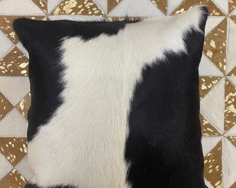 May include: Black and white cowhide pillow with a soft, fluffy texture. The pillow is shown on a brown and white geometric patterned rug.