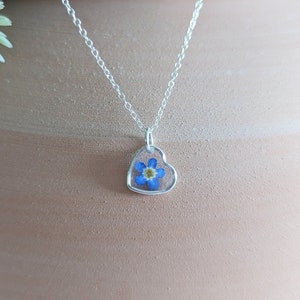 May include: A silver chain necklace with a heart-shaped pendant featuring a pressed blue forget-me-not flower.