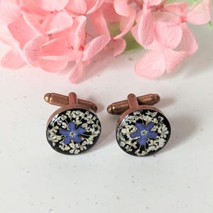 May include: A pair of cufflinks with a copper finish. Each cufflink features a round, black resin base with a single blue flower surrounded by white flowers pressed into the resin.