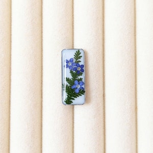 May include: A small rectangular resin charm with a white background and a pressed blue flower design.