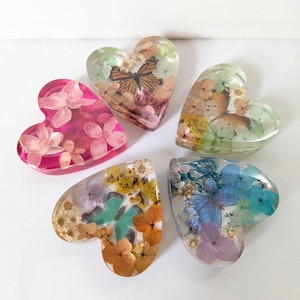 May include: Five heart-shaped resin paperweights containing preserved flowers and butterfly wings. One is pink with pink flowers, while the others are clear, featuring various combinations of yellow, blue, purple, and orange flowers alongside butterfly wings.