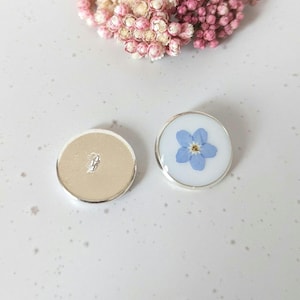 May include: Two round, silver-rimmed buttons with a white background. One button features a pressed forget-me-not flower in blue.