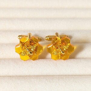May include: A pair of gold cufflinks with a honeycomb design and a bee in the center. The cufflinks are made of a clear, amber-colored material.