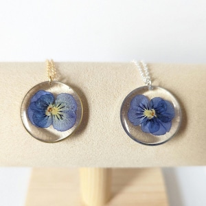 May include: Two necklaces with round pendants featuring pressed purple pansies. One necklace has a gold chain and the other has a silver chain.