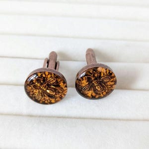 May include: A pair of gold and brown cufflinks with a bee design. The cufflinks are on a white background.