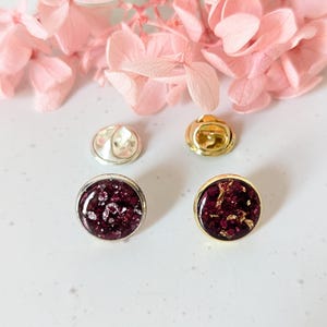 May include: Two round pins with a dark purple resin base and gold or silver accents. The resin has a glittery finish. The pins are on a white surface.