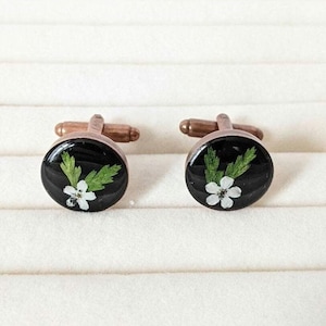 May include: A pair of cufflinks with a black background and a white flower with green leaves pressed inside.