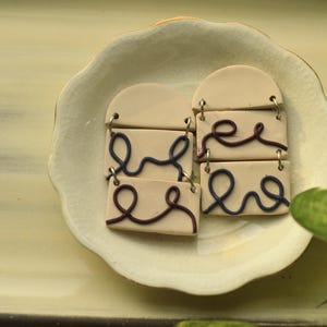 Könnte beinhalten: Zwei Paar Keramikohrringe mit weißer Basis und einem Design aus schwarzen und blauen Linien. Die Ohrringe befinden sich auf einem weißen Teller.