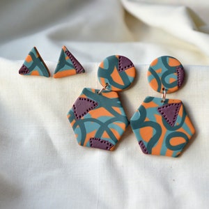 May include: A set of three pairs of earrings featuring abstract geometric designs in orange, teal, and purple. The earrings are made of polymer clay and have a modern, abstract style.