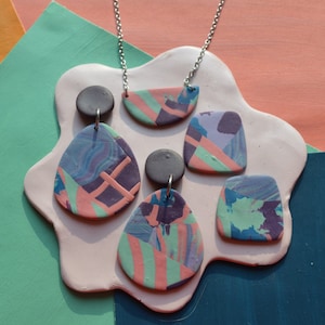 May include: A set of colorful polymer clay earrings and a necklace. The earrings are shaped like ovals and the necklace is a crescent shape. The colors are pink, green, purple, and blue.