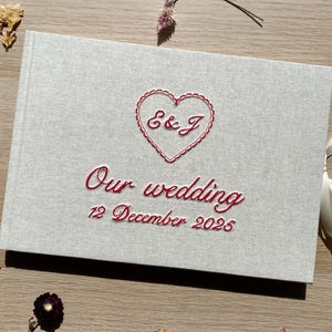 May include: A light gray wedding guest book with red embroidered text and a heart design. The text reads "E & J" inside the heart, and "Our wedding 12 December 2025" below. A cream ribbon is tied to the side.
