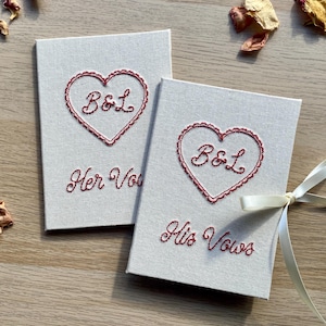 May include: Two off-white vow books with red embroidered heart designs and the initials "B&L". One book reads "Her Vows" and the other "His Vows". A cream ribbon ties the books together. Dried flower petals are scattered around.