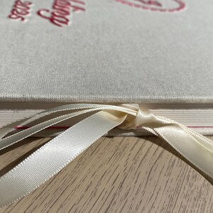 May include: Close-up of a cream-colored book with the word "Catalog" and the year "2025" embossed in red. A cream-colored ribbon is tied around the book. The book is on a light wood surface.