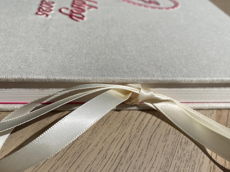 May include: Close-up of a cream-colored book with the word "Catalog" and the year "2025" embossed in red. A cream-colored ribbon is tied around the book. The book is on a light wood surface.