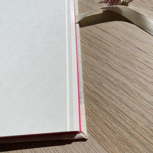 May include: A hardcover book with white pages and a pink edge. The book has a light-colored linen cover and a cream-colored ribbon bookmark. A small dried flower rests on the wooden surface.