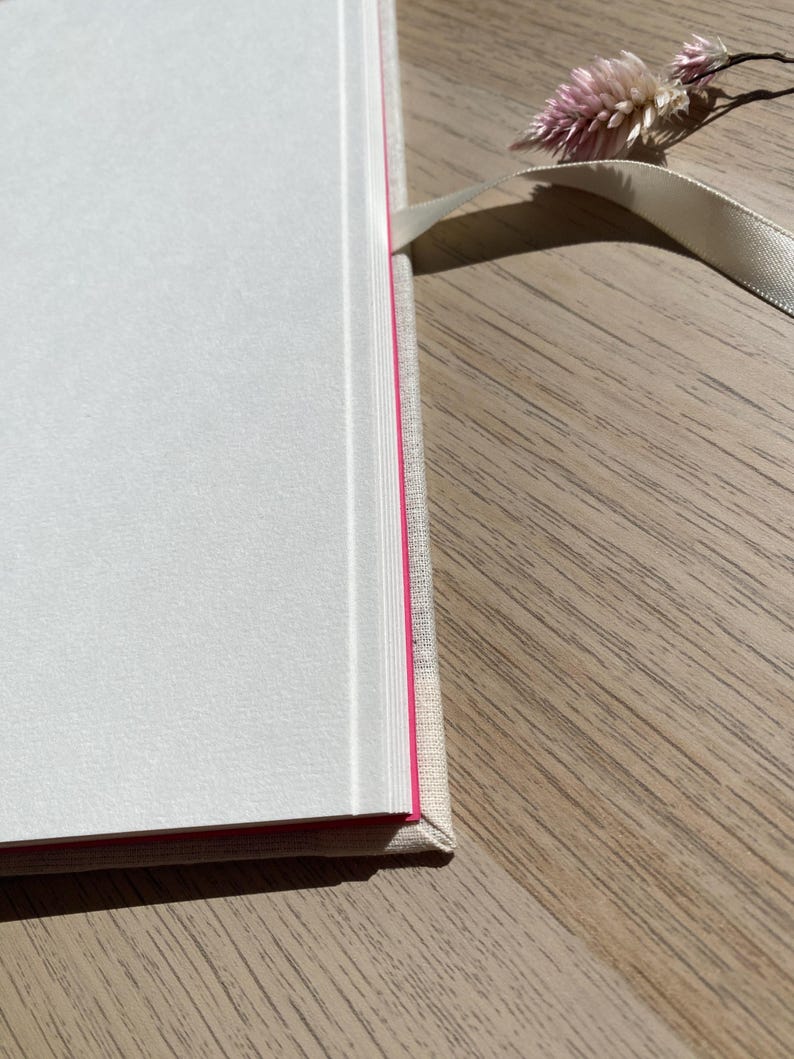 May include: A hardcover book with white pages and a pink edge. The book has a light-colored linen cover and a cream-colored ribbon bookmark. A small dried flower rests on the wooden surface.