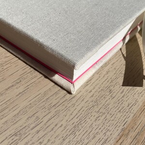 May include: A close-up of a hardcover book with a light beige linen cover. The book's edges are white with a thin pink line. A cream-colored ribbon is attached. The book is on a light brown wooden surface.