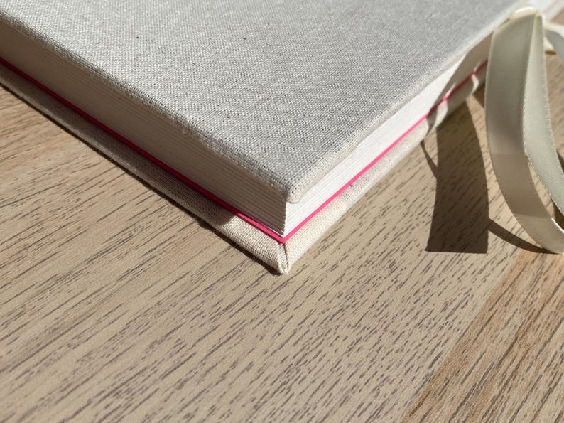 May include: A close-up of a hardcover book with a light beige linen cover. The book's edges are white with a thin pink line. A cream-colored ribbon is attached. The book is on a light brown wooden surface.