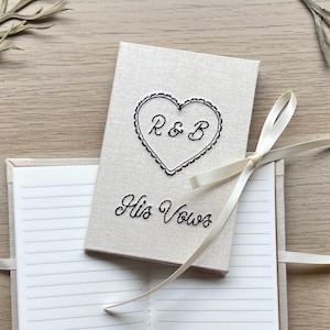 May include: A beige linen-textured wedding vow book with a black embroidered heart design containing the letters "R & B". The words "His Vows" are embroidered below the heart. A cream-coloured ribbon is tied around the book.