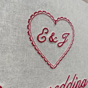 May include: Close-up of a linen fabric with red embroidery. A heart shape with scalloped edges contains the letters "E & J". Below the heart, the word "wedding" is embroidered in a cursive font. The fabric is a light gray color.
