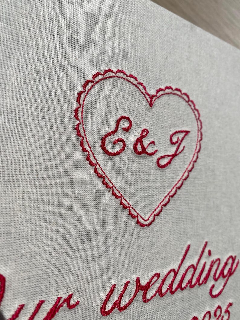 May include: Close-up of a linen fabric with red embroidery. A heart shape with scalloped edges contains the letters "E & J". Below the heart, the word "wedding" is embroidered in a cursive font. The fabric is a light gray color.