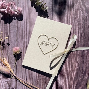 Könnte beinhalten: Eine cremefarbene Hochzeitskarte mit einem goldgestickten Herz und den Initialen "J&M". Die Karte ist mit einem cremefarbenen Band gebunden und liegt auf einer Holzoberfläche, umgeben von getrockneten Blumen und Blättern.