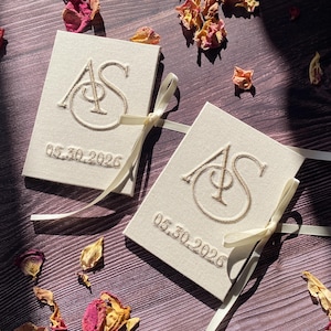 Pode incluir: Duas caixas de lembrancinhas de casamento em creme com as iniciais bordadas "AS" e a data "05.30.2026". Cada caixa é amarrada com uma fita creme. Pétalas de rosa secas espalhadas em uma superfície de madeira.