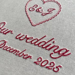 May include: Close-up of a light gray fabric with red embroidered text and a heart. The heart contains the initials "E & J". Below the heart, the text reads "Our wedding December 2025".
