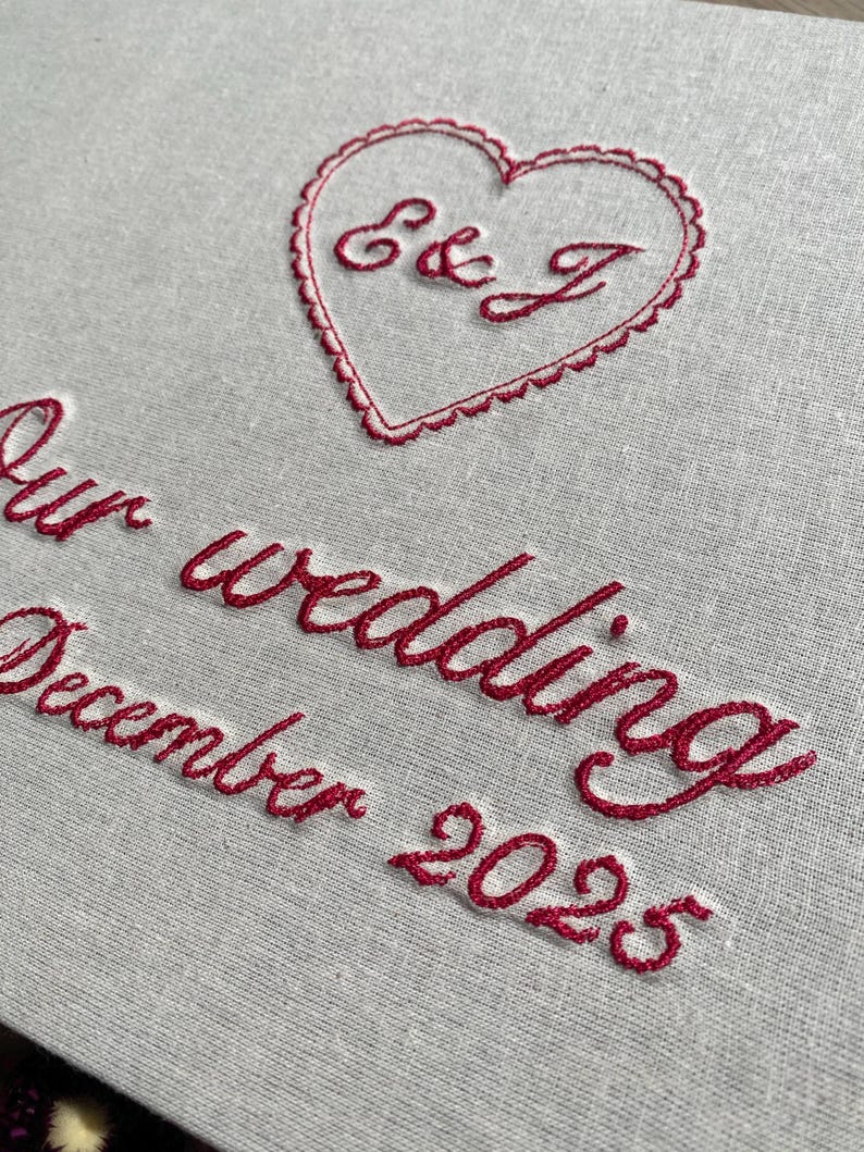 May include: Close-up of a light gray fabric with red embroidered text and a heart. The heart contains the initials "E & J". Below the heart, the text reads "Our wedding December 2025".
