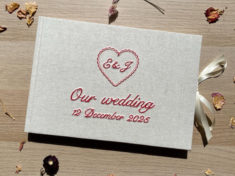 May include: A light gray wedding guest book with red embroidered text and a heart design. The text reads "E & J" inside the heart, and "Our wedding 12 December 2025" below. A cream ribbon is tied to the side.