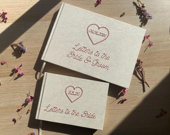 May include: Two beige fabric-covered books with embroidered pink text and heart designs. The larger book reads Letters to the Bride & Groom with 06.06.2026 inside a heart. The smaller book reads Letters to the Bride with 9.5.26 inside a heart.